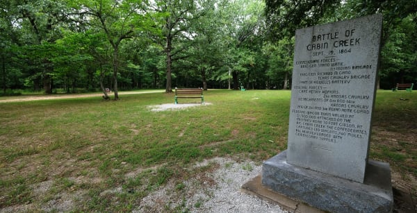 The granite Battle of Cabin Creek marker beside a gravel path in a mown, tree-shaded field at the battlefield near Pensacola.