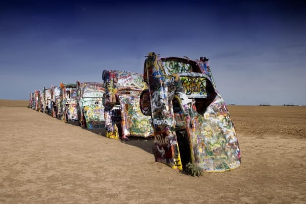 A row of graffiti-covered Cadillacs buried nose down in bare dirt at Cadillac Ranch under a deep blue sky.