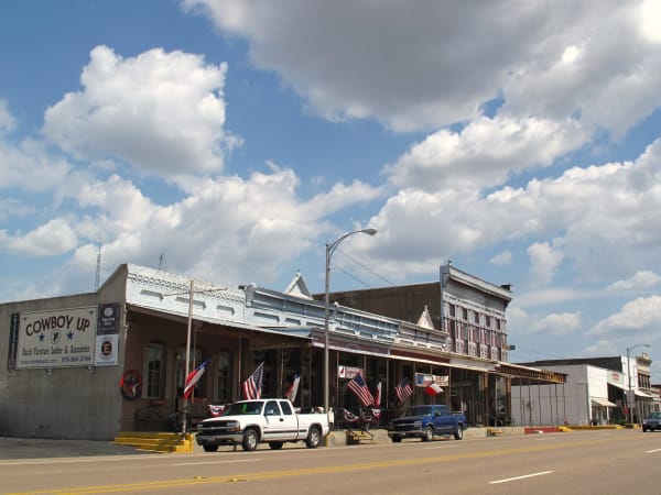 Main Street in Calvert, with brick storefronts and a two-story building with a long porch under a cloud-dotted sky.