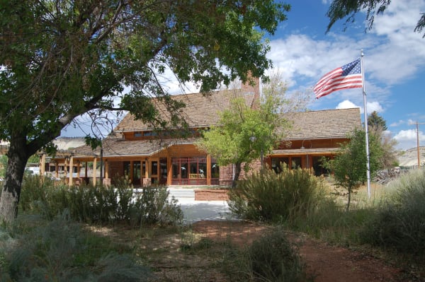A single-storey visitor centre at Cannonville — a broad shingled roof over a deep porch carried on log posts and red sandstone piers, glazed doors with bird decals set between them, and a US flag flying from a white pole at right. Cottonwoods frame the view, sagebrush and rabbitbrush fill the foreground beside a concrete approach path, and pale bluffs show behind under blue sky and cumulus.