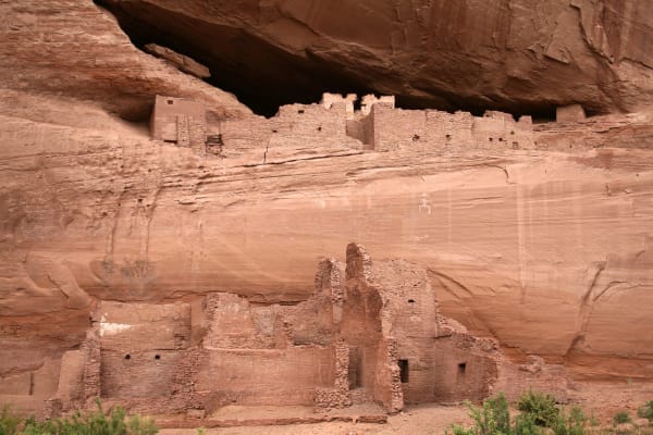 White House Ruin in Canyon de Chelly — Tséyiʼ — an Ancestral Puebloan village built against a sheer sandstone wall on Diné land. The lower block stands on the canyon floor behind a screen of brush, its coursed masonry rooms broken open to show square doorways and standing corners; the upper block sits in a shallow alcove under a great overhang, its rooms stepped along the ledge behind a low parapet with a pale plastered wall at the left end. A panel of white pictographs runs across the cliff face between the two levels, and the sandstone above is streaked with desert varnish.