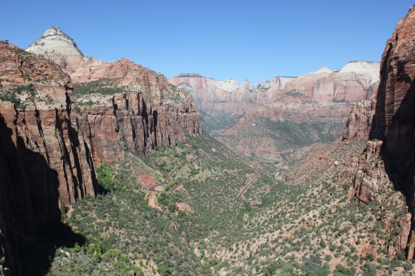 The view from the Canyon Overlook — a deep canyon dropping away between sheer red walls and fluted buttresses, the highway switchbacking up its floor in tight hairpins through pinyon and juniper, and white-capped sandstone peaks standing across the canyon mouth in the distance under a cloudless sky.