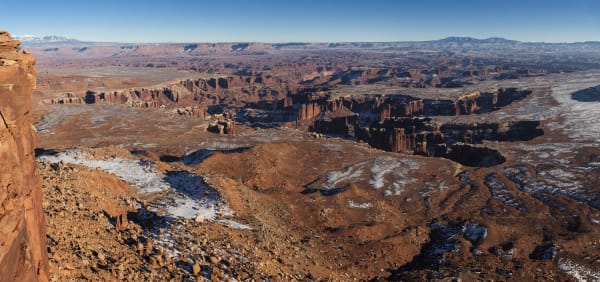A wide panorama from a canyon rim in winter, looking out over tiers of red rock benches and buttes with snow lying in the hollows. A dark cliff edge cuts into the frame at the left, and distant snow-capped mountains sit on the horizon under a clear sky.