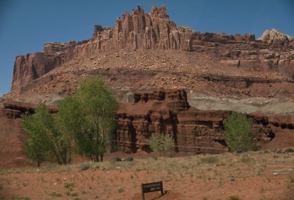 The Castle in Capitol Reef — a wall of dark red sandstone rising to a crown of ragged spires and turrets above a long scree apron, with lower banded ledges and grey-green shale slopes stepping down to a flat of red soil and sparse brush. A brown park sign reading CAMPGROUND with an arrow stands in the foreground and a highway sign for Hanksville and Torrey at the right, under a deep blue sky.