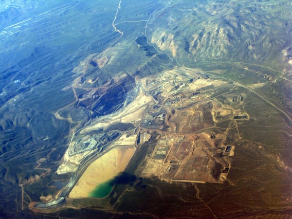 An oblique aerial photograph of a large open-pit gold mining complex in northern Nevada — benched pits cut into the hillsides, pale waste rock dumps and haul roads spreading across the ground, process buildings and rectangular leach pads at the right, and a tailings impoundment at the bottom with a tan beach and a green pond. Hazy blue-grey ranges run away to the top of the frame.