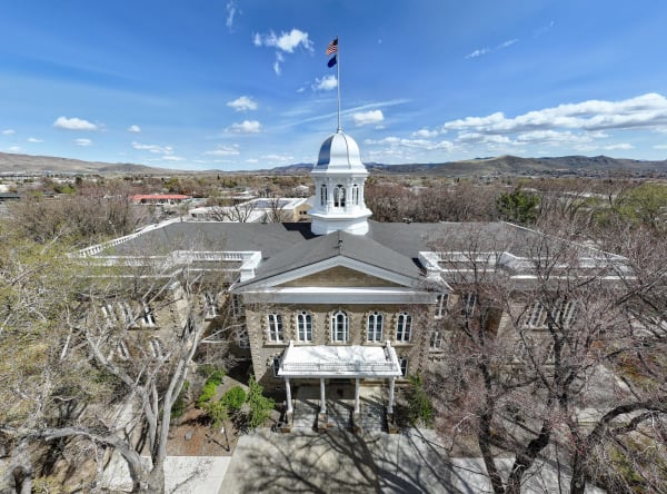 An elevated view of the Nevada State Capitol in Carson City, its sandstone walls topped by a white cupola and silver dome flying the U.S. and Nevada flags, bare early-spring trees around the grounds and low hills beyond.