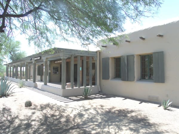 The visitor center at Casa Grande Ruins National Monument — a low pale stucco building with a shallow parapet, deep-set windows in dark green frames, and a timber ramada on square posts running the length of its front, with VISITOR CENTER and NATIONAL PARK SERVICE signs and a notice case in the shade beneath it. Agaves and a barrel cactus grow in the gravel forecourt, a mesquite overhangs the frame from the upper left and throws its shadow across the paving. The Casa Grande itself — the four-storey caliche great house the Huhugam raised, and the steel canopy standing over it — is not in the frame.