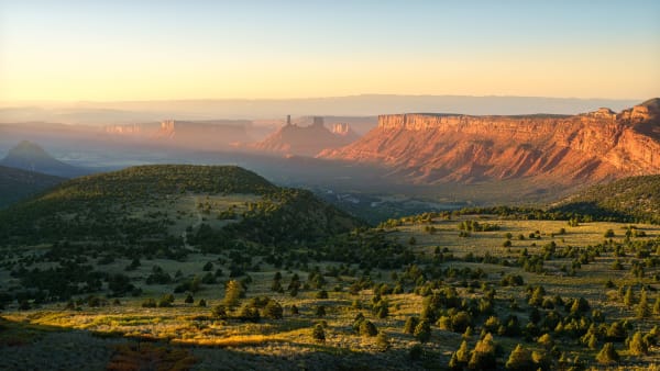 A broad valley at sunrise seen from above, juniper-dotted grassland in the foreground running to a long red mesa wall lit gold along its rim on the right. A slender rock tower and smaller spires rise from the valley floor in the middle distance, with hazy layered cliffs beyond.