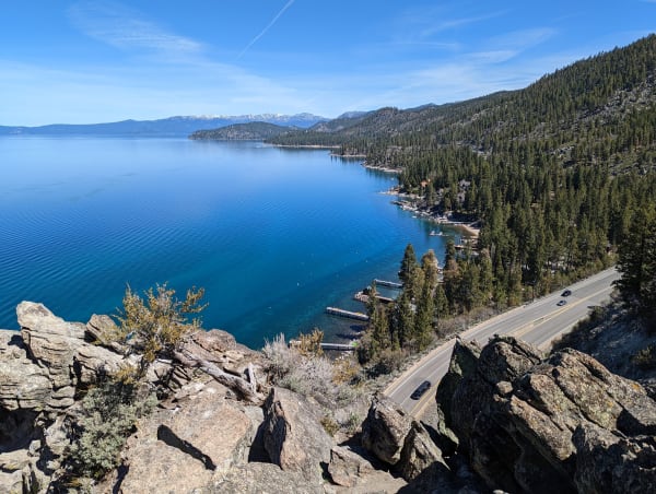 The view from the rocky top of Cave Rock over Lake Tahoe, U.S. 50 curving along the shoreline below past small piers, forested slopes rising to the right and snow-flecked peaks on the far side of the lake.