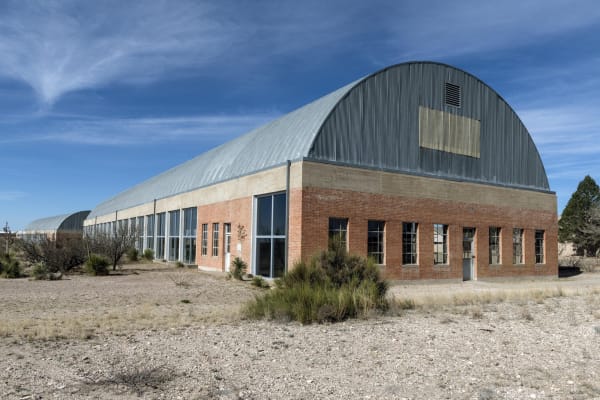 The arched metal roof and long brick walls of the Chinati Foundation headquarters building in Marfa, set among desert grasses.