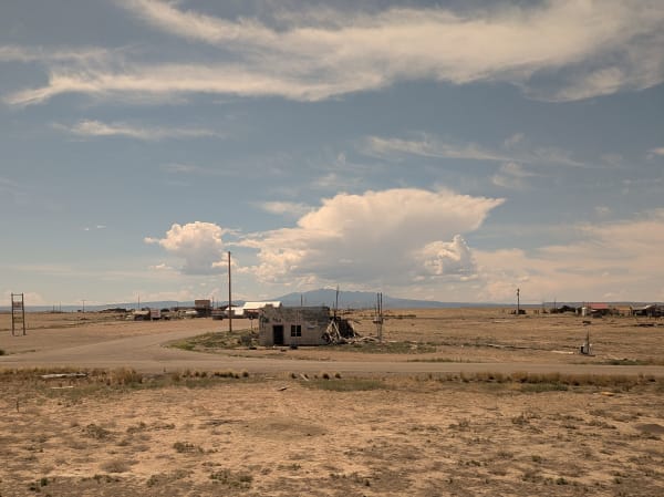 A wrecked single-story stone building with a collapsed roof standing alone beside a paved road on flat desert, scattered sheds, poles and derelict vehicles spread thinly behind it. A tall cumulus cloud rises over low distant mountains and dry grass fills the foreground.