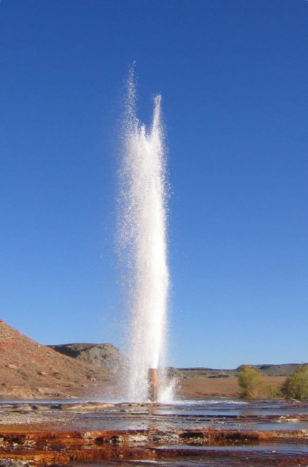 A tall narrow column of white water erupting from a low pipe at ground level and breaking into spray at its top, against a cloudless deep blue sky. Rust-orange mineral terraces and shallow pools spread across the foreground, with low desert hills on both sides.