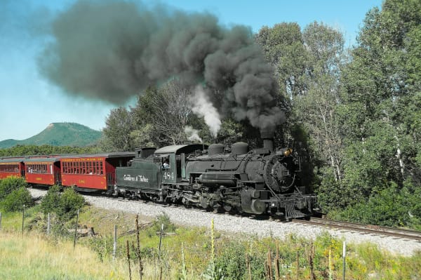 A Cumbres and Toltec steam locomotive trailing black smoke as it pulls red passenger coaches past green trees