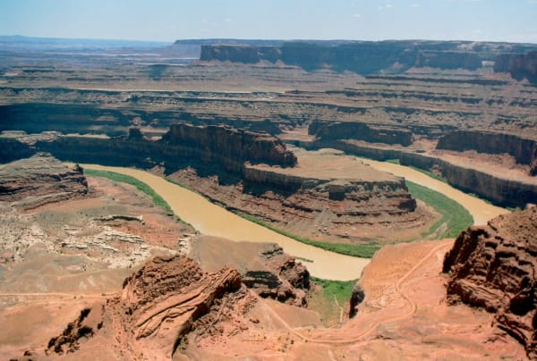A tight loop of the Colorado River seen from high on the canyon rim, the pale tan water curving around a green-fringed bench of red rock. Layered cliffs and benches step away in every direction to flat-topped mesas on the hazy horizon.