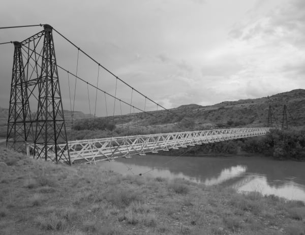 An archival black and white photograph of the suspension bridge complete and intact, its timber deck and trussed railings carried on cables from a tall steel tower at the left, spanning the Colorado River. Dry grass fills the foreground and low desert hills rise beyond under an overcast sky.