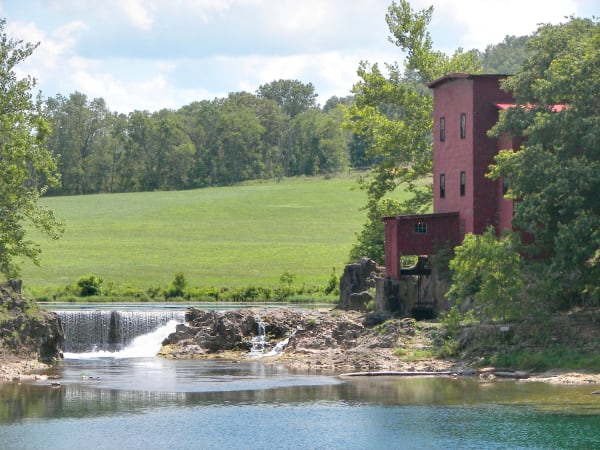 The red frame Dillard Mill beside the rock dam and millpond on Huzzah Creek, with green fields behind.