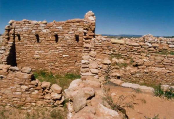 Excavated Ancestral Puebloan masonry at the Edge of the Cedars pueblo in Blanding — a standing block of coursed sandstone at the left with a row of four small square openings set high in the wall and a dark doorway at its near corner, built of thin flat slabs laid in tight courses with chinking stones packed between them. Lower stabilised walls run away to the right across cropped green grass and red earth, fallen blocks and a slab-paved surface lie in the foreground, and a low blue ridge stands on the far horizon under a hard clear sky. The kiva and the ladder down into it are not in the frame.