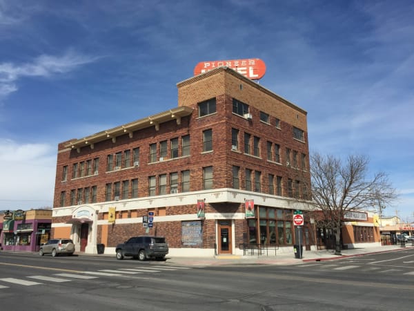 The Pioneer Hotel in Elko seen from below at its corner — four storeys of red and tan brick with a stepped parapet and window air conditioners in several openings, and a large red oval roof sign reading PIONEER HOTEL with much of its lettering missing. A bare tree stands at the right against a blue sky with thin cloud.
