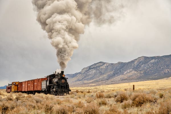 Nevada Northern Railway locomotive No. 40 under steam on the open flats below a limestone range, a tall pale plume standing straight up from the stack. Behind the tender run three oxide-red wooden boxcars, a yellow bay-window caboose and a dark red coach, with winter sagebrush filling the foreground and a crew member sitting in the cab doorway. The town of Ely is not in the frame.
