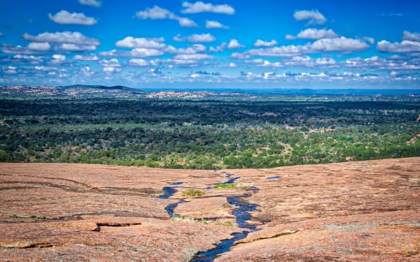Bare pink granite with rainwater pools on top of Enchanted Rock, looking out over wooded Hill Country under scattered white clouds.