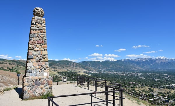 The cobblestone obelisk on the summit of Ensign Peak, standing on a paved terrace behind pipe railings with an interpretive plaque beside it, and the Salt Lake Valley spread below — downtown towers among green neighbourhoods, with the snow-streaked Wasatch Range across the valley under a clear blue summer sky.
