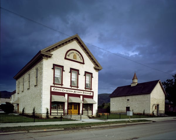 A two-story building of pale cream limestone seen from across the street under a heavy purple storm sky. Ephraim Co-operative Mercantile Association is lettered across the front above striped awnings and a yellow door, and a beehive with the words Holiness to the Lord fills the gable. A second stone building with a small cupola stands to the right.