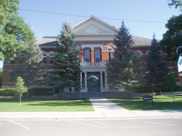 A broad red brick college building seen head-on from a parking lot, its white pedimented gable carrying a carved crest and SNOW COLLEGE cut into the stone above an arched entrance porch with white columns. Blue spruces stand along the front and a lawn runs down to the street.