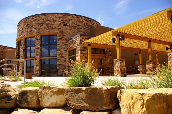 The Escalante Interagency Visitor Center — a drum-shaped rotunda faced in thin stacked sandstone with tall arched windows, a stuccoed wing beyond carrying a sign reading VISITOR CENTER above glazed doors, and a long timber trellis walkway on stone piers running away to the right. A boulder retaining wall, grasses and orange-flowered shrubs fill the foreground, under blue sky streaked with thin cloud.