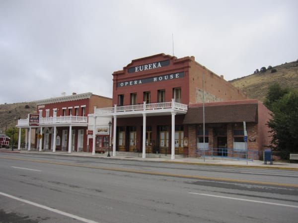 The Eureka Opera House on US-50 — a two-storey red brick building with EUREKA OPERA HOUSE lettered in white across its parapet and a white-columned porch and balcony along its front. The Jackson House Hotel, Saloon & Cafe adjoins it on the left and a Keyhole Bar sign hangs between them; the highway's yellow centre line and empty asphalt cross the foreground, with a dry juniper-dotted hillside behind.