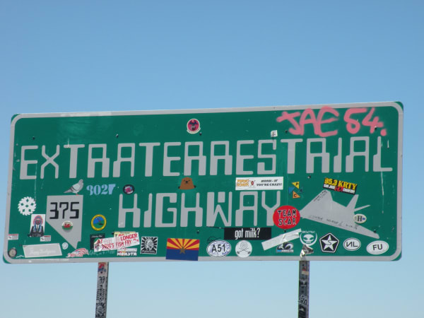 The Extraterrestrial Highway sign for Nevada State Route 375, filling the frame against a clear sky — a wide green sign with EXTRATERRESTRIAL HIGHWAY in white block letters, a route 375 shield set in the outline of Nevada at the lower left, and JAE 54 sprayed across the top right corner in pink. The lower half is layered with travellers' stickers and marker graffiti: an Arizona state flag, a hand-drawn grey flying wing, an alien head, radio-station and band decals, joke slogans and dozens of smaller labels. Both steel posts are wrapped in stickers down to the bottom edge, and nothing else stands in the frame.