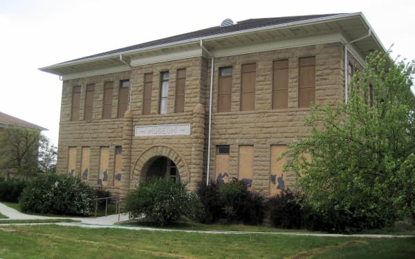 A two-story building of rough faced tan sandstone photographed from below at its corner, MUSEUM lettered on a panel above a round arched doorway. Dark metal silhouettes of people and animals are mounted along the lower wall, shrubs crowd the base, and the sky above is flat white overcast.