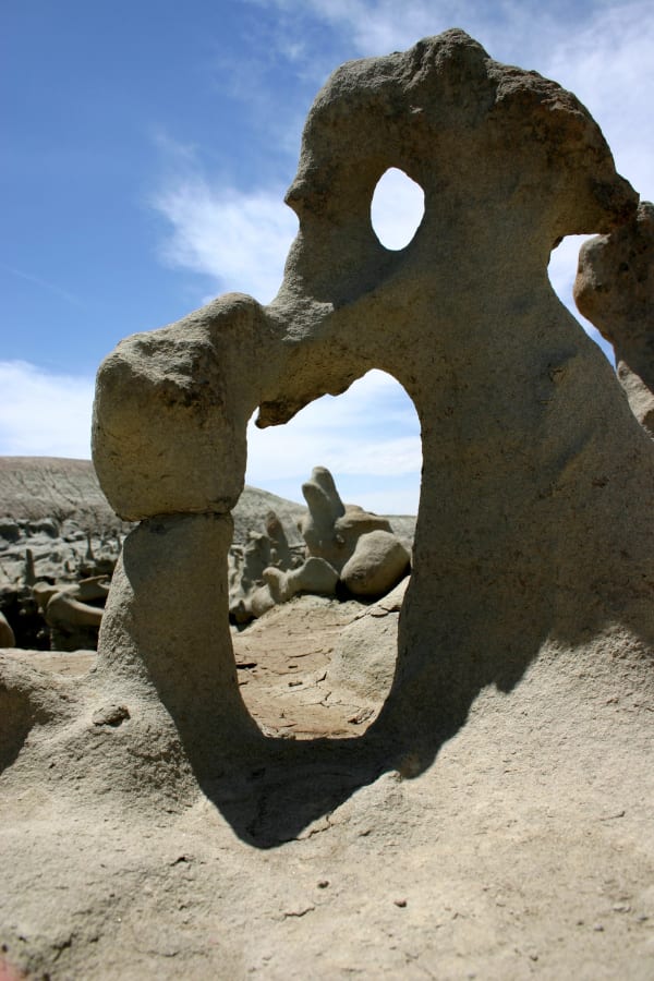 A single knobbed sandstone formation at Fantasy Canyon photographed from ground level against a blue sky — a tall pierced fin with a wide window through its base and a smaller round hole higher up, its whole surface eroded into lumps and lobes. More low formations and a cracked mud floor show through the opening, with pale badland ridges beyond.