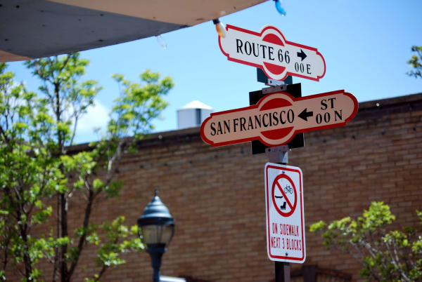 The Route 66 and San Francisco Street corner sign in downtown Flagstaff, shot close from below — two enamelled blade signs crossed on a single post, the upper lettered ROUTE 66 with a right arrow and 00 E, the lower SAN FRANCISCO ST with a left arrow and 00 N, both cream on red with a round Route 66 shield at each end. A regulatory sign beneath them prohibits cycling on the sidewalk for the next three blocks. A red brick wall, a green tree and the edge of a white awning fill the background. The town itself, the ponderosa forest and the observatory are all out of frame.