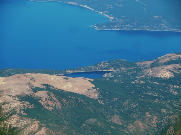 A hazy aerial view over the forested Carson Range, with dark blue Marlette Lake held in a basin on the ridge and the far larger expanse of Lake Tahoe beyond it, a developed shoreline visible across the water.