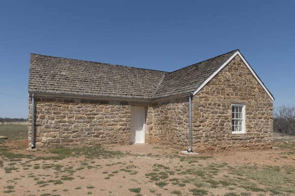 The restored post bakery at Fort Griffin, a one-story sandstone building with a wood-shingle roof, standing on bare red ground under a clear sky.