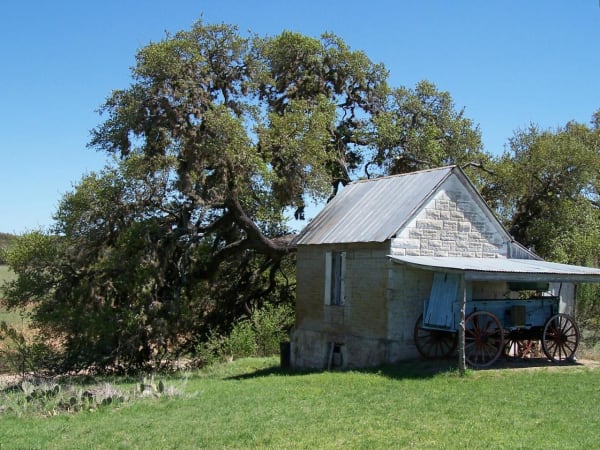 A small stone building with a tin roof and an old wagon under its shed, beside a large oak at Fort Martin Scott.