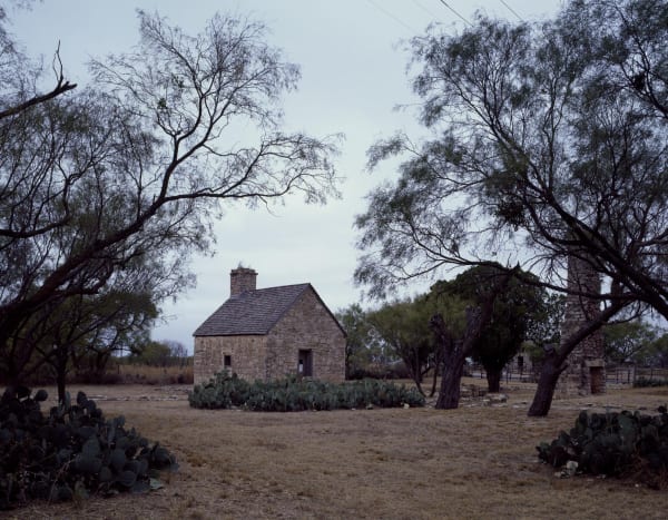 A small stone building with a single chimney among mesquite trees and prickly pear at Fort Phantom Hill, with a lone stone chimney standing at right.
