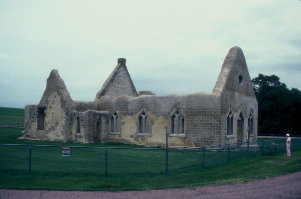 The roofless chalkstone walls and gables of the 1875 Fort Randall chapel, behind a low fence on the grass