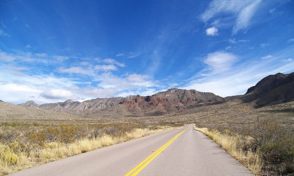 A two-lane road running through desert scrub toward the rugged peaks of the Franklin Mountains in El Paso.