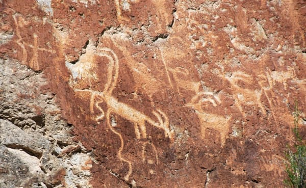Petroglyphs pecked into a red rock face, the pale figures standing out where the darker surface has been cut away. Several horned four-legged animals cross the panel, a long wavy line runs down the left side, and a human figure with outstretched arms and legs stands at the upper left.
