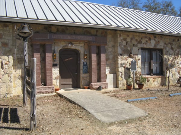 The stone front of the Frontier Times Museum in Bandera, with a wooden door, an old bell on a post and a ramp to the entrance.