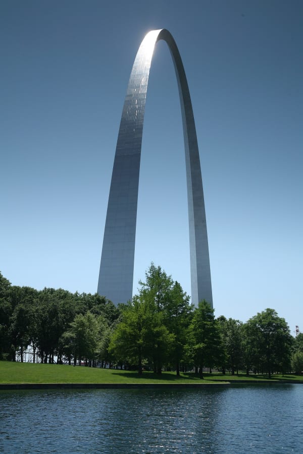 The stainless steel Gateway Arch rising above a line of trees and reflected in a park pond.
