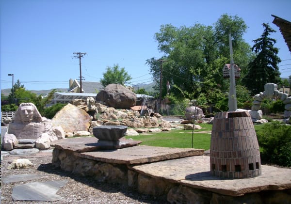 Carved stones set across the lawn at Gilgal Sculpture Garden — a sphinx with a human face at left, a brick beehive-shaped tower at right, and a stone basin resting on flagstone slabs in the foreground, with neighbouring rooftops, power lines and the foothills rising behind.