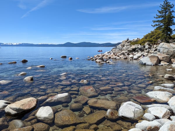 Clear shallow water over rounded granite boulders on Lake Tahoe's east shore, a low rocky point running out to the right with two people sitting on it, a red kayak offshore and snow-streaked peaks along the far shoreline.