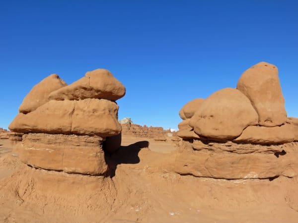 Goblins at Goblin Valley seen close and from below — squat orange sandstone knobs with rounded, top-heavy caps balanced on narrower pedestals, ranged along banded ledges on both sides of the frame with more of them running away in ranks through the gap between, under a cloudless deep blue sky.