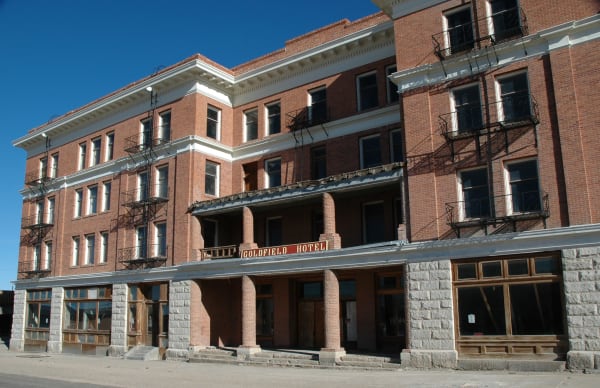 The Goldfield Hotel on its corner, four storeys of red brick above a rusticated grey stone ground floor, with a painted GOLDFIELD HOTEL board hung beneath the first-floor canopy and iron fire escapes zigzagging down the facade. Rows of tall windows stand blank or open to the dark, the ground-floor shopfronts are boarded behind their glass, bare concrete steps run up to the doorways, and empty pavement crosses the foreground under a hard blue sky.