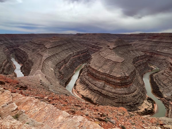 The entrenched meanders of the San Juan River seen from the rim at Goosenecks State Park, the river doubling back on itself around long grey fins of layered rock a thousand feet below. Pale green water catches the light at the left, again in the centre and again at the right, each bend divided from the next by a narrow spine of banded shale and limestone stepping down in hundreds of thin ledges. Broken red slickrock and loose rubble fill the foreground at the edge of the overlook, and a heavy grey storm lies low across the whole canyon.