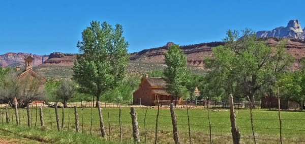 Grafton across a hayfield — the brick schoolhouse with its white bell cupola at left and a two-storey brick house with a covered porch at centre, both standing among cottonwoods behind a leaning barbed-wire fence on weathered posts. Red and grey benchland rises behind, with a white sandstone peak at the far right under deep blue sky.