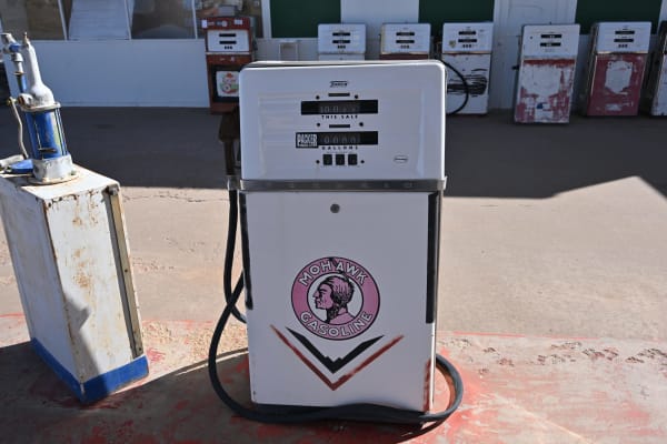 A restored Tokheim gasoline pump standing on a concrete forecourt at Grand Canyon Caverns, filling the frame — white with a chrome band, its dial faces lettered THIS SALE and GALLONS, a black hose looped to the ground, and a Mohawk Gasoline brand disc on its lower door carrying a mid-century Native American head device. A rusted white service cabinet stands at the left, and a row of seven more vintage pumps in white, red and cream is ranged along a building behind, low sun throwing their shadows across the apron. The cavern itself, and the 210-foot descent to it, are not in the frame.