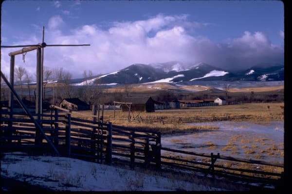 Pole corrals and a loading chute at Grant-Kohrs Ranch, wet meadow and ranch buildings beyond, snow on the mountains behind